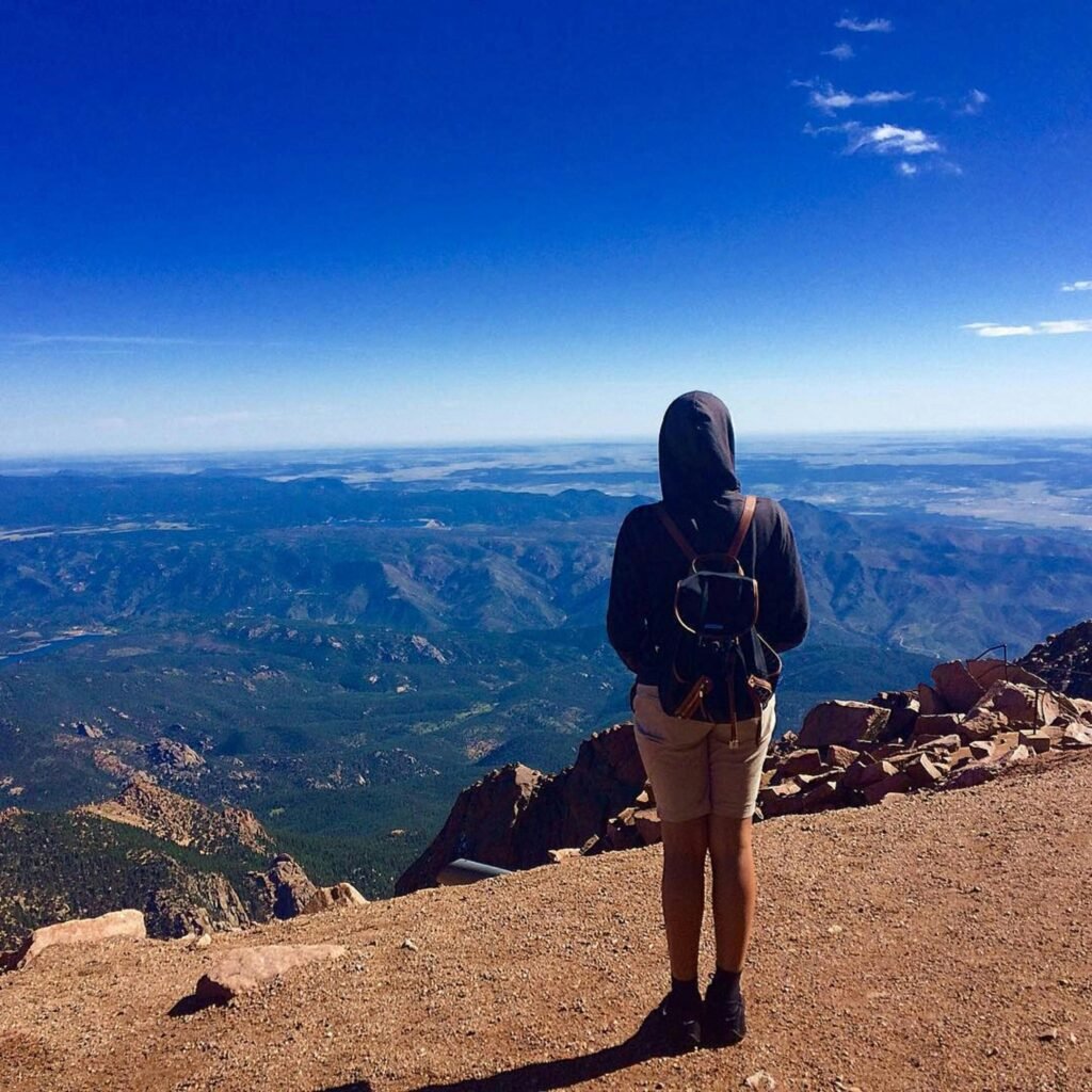 A solo hiker with a backpack stands atop Pikes Peak in Colorado, taking in a stunning mountain view.
